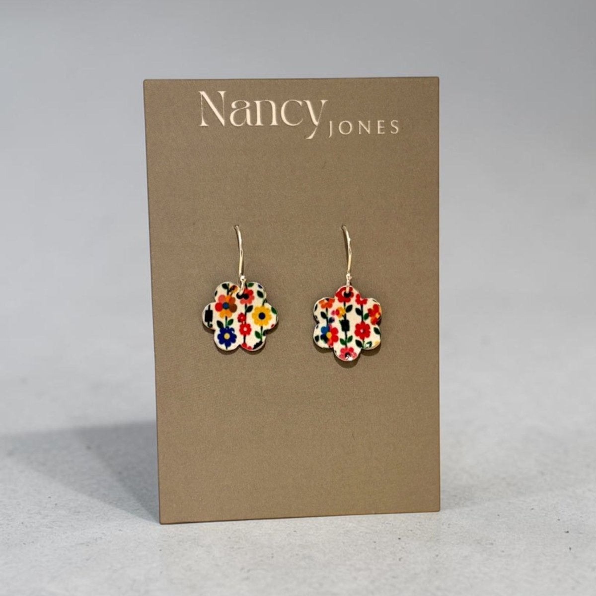 A pair of Nancy Jones floral enamel flower-shaped drop earrings displayed on a brown cardboard brand card against a neutral gray background.