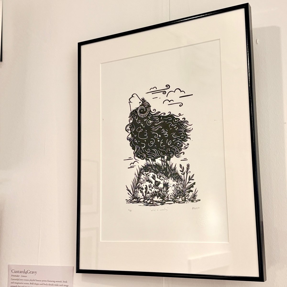 A framed black and white art print featuring a whimsical sheep illustration, displayed on a white gallery wall with a small label card visible at the bottom left.