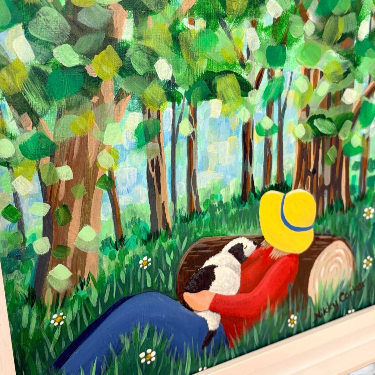 A colorful painted scene of a person in a red shirt and yellow hat lying on a log in a forest, holding a black-and-white dog. The artwork fills most of the frame, with a white wall or surface visible around the edges.