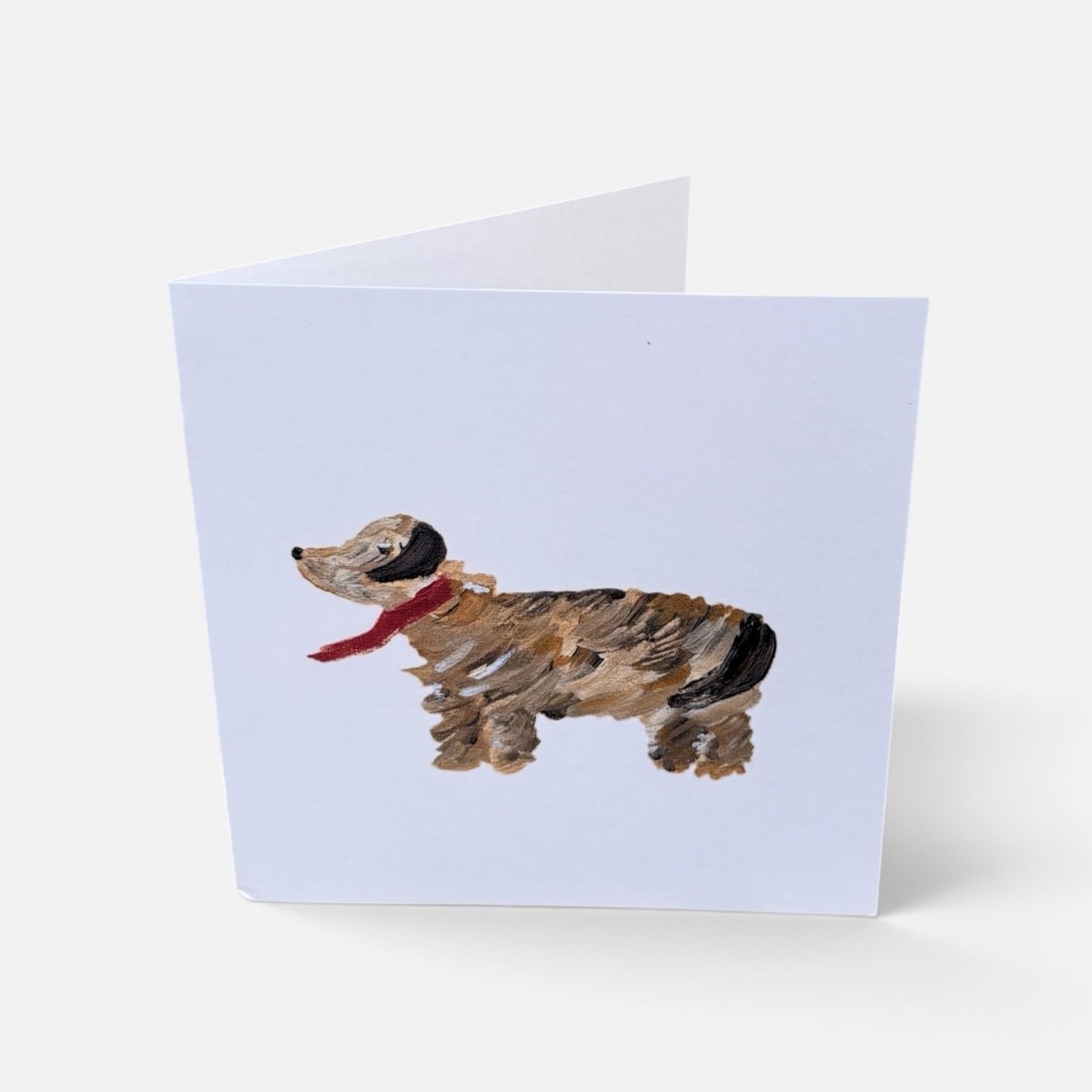 A folded greeting card standing upright on a light gray background. The front of the card features a painted dachshund in brown, black, and tan with a red scarf on a pale lavender card background.