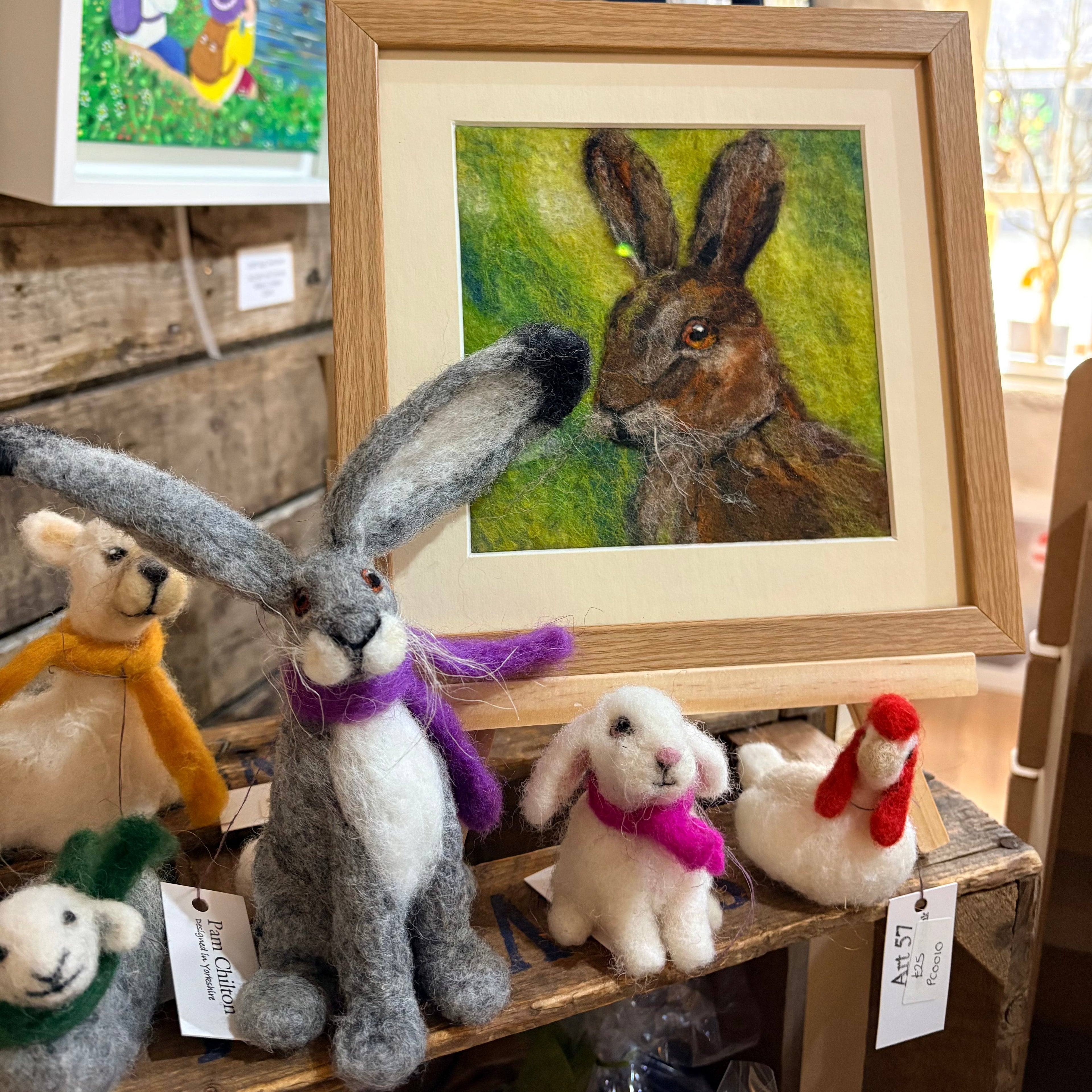 Felted animals on a wooden shelf with a framed artwork of a hare.
