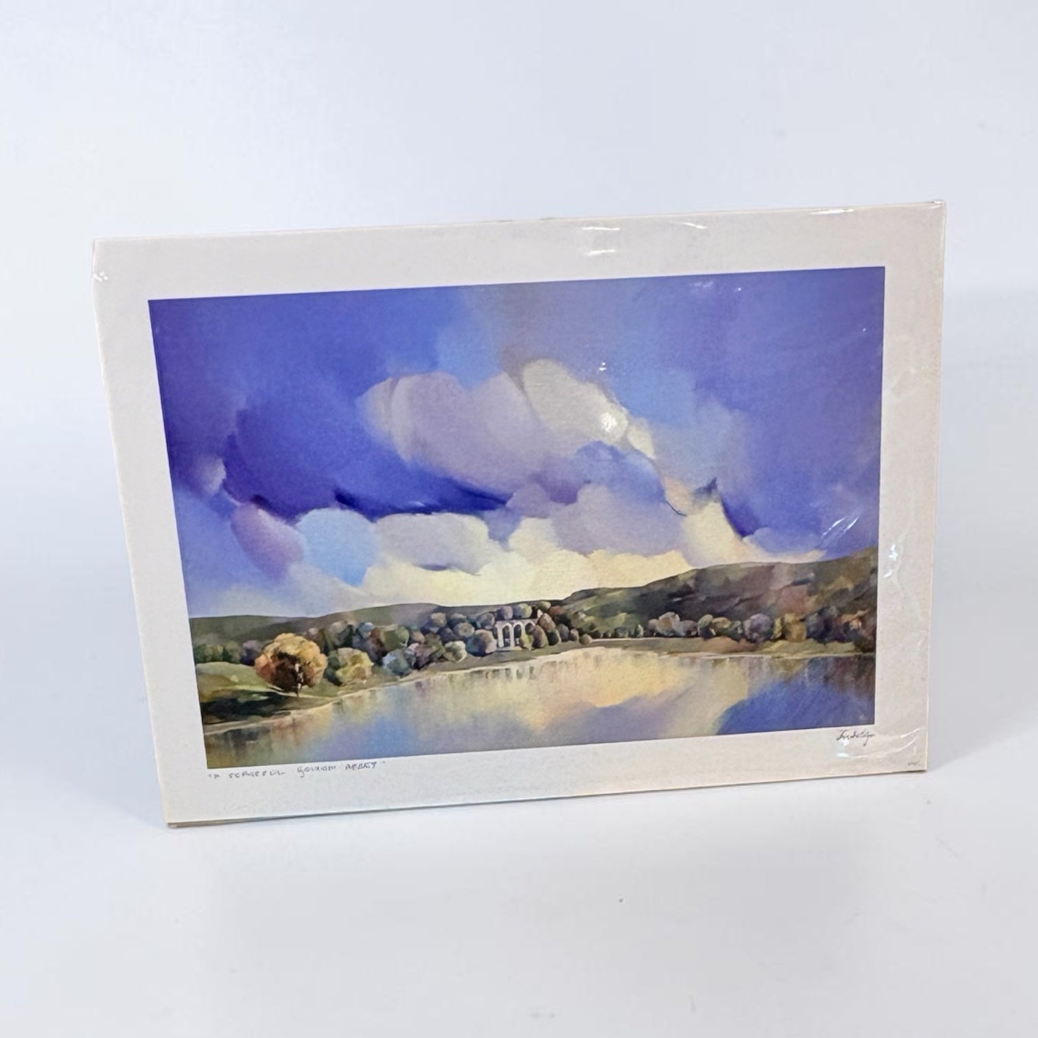 A square watercolor landscape art print in its original white paper border and plastic wrapping, showing a blue and purple sky over a reflective lake with trees and hills.