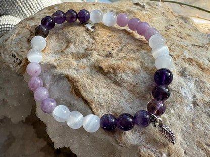 A beaded stretch bracelet made of alternating translucent white, lavender purple, and deep amethyst beads, with a small silver feather charm, displayed on a natural rock surface.