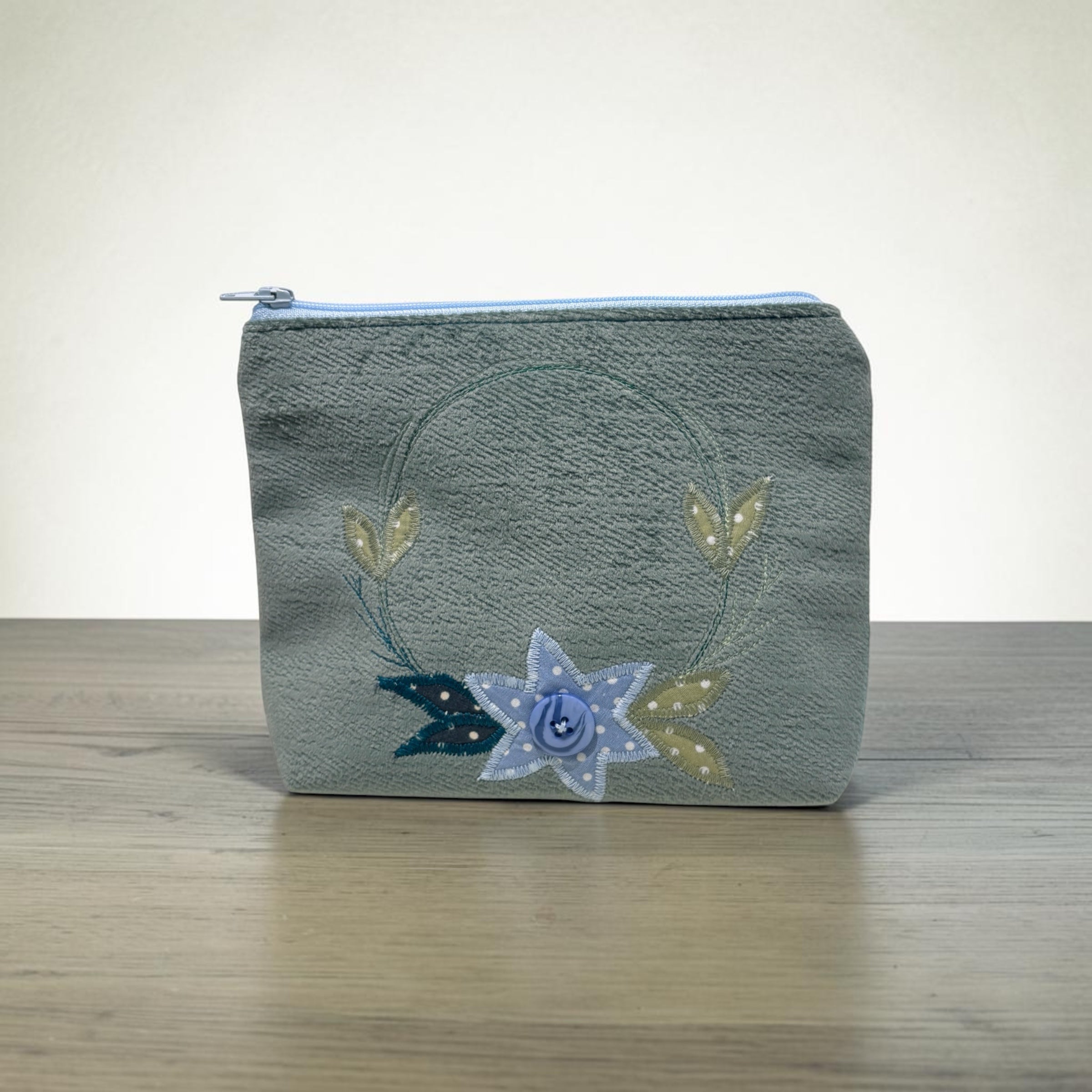 A small zippered pouch with a textured gray-green fabric body, featuring a decorative embroidered design of a blue and white star, gold leaves, and a blue ribbon on a light wood surface against a neutral background.
