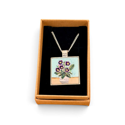 A square pendant necklace displayed inside an open tan gift box with a black insert. The pendant shows a floral bouquet in a vase with purple flowers and green leaves on a light blue and beige background, suspended from a silver-toned chain.