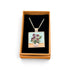 A square pendant necklace displayed inside an open tan gift box with a black insert. The pendant shows a floral bouquet in a vase with purple flowers and green leaves on a light blue and beige background, suspended from a silver-toned chain.