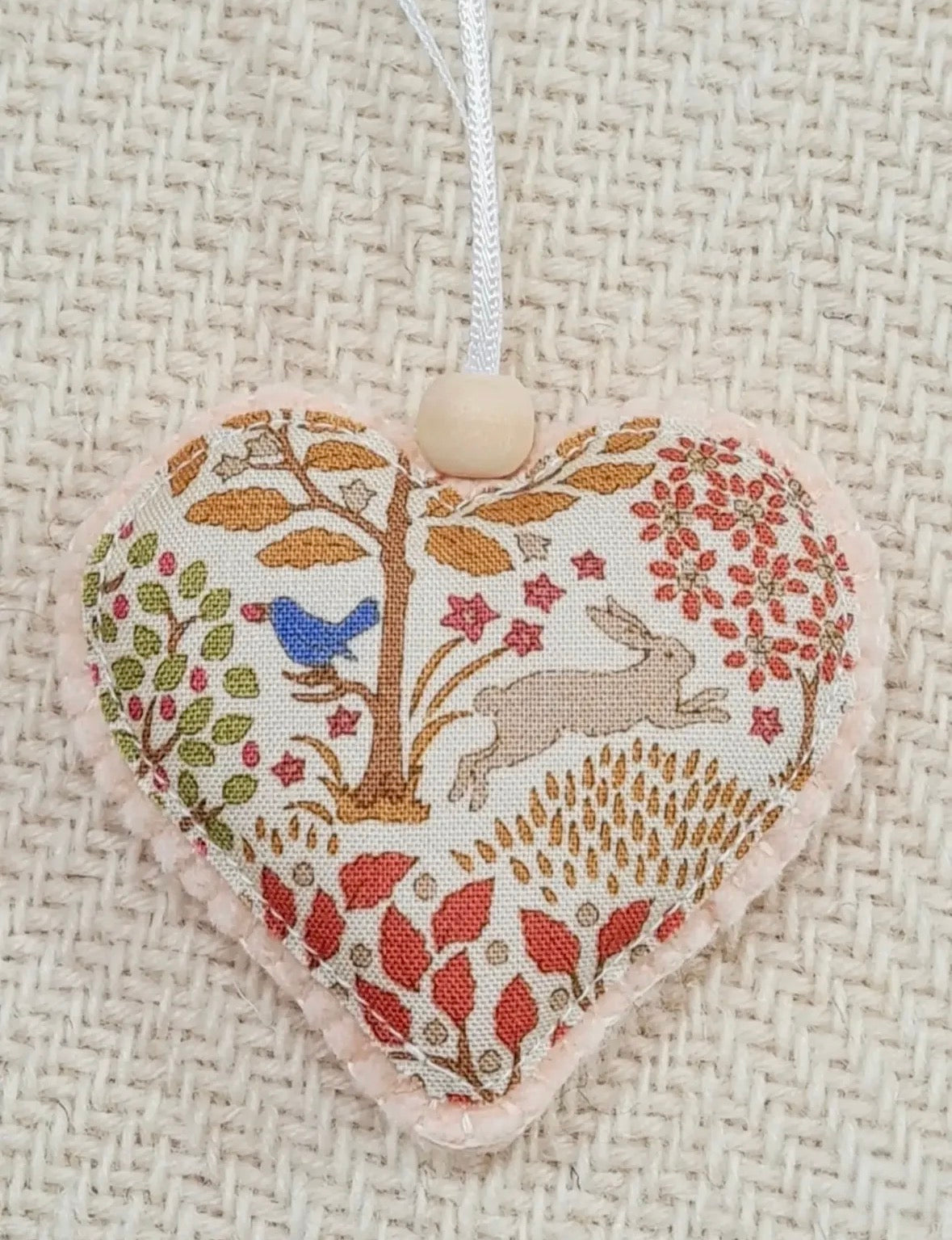 A heart-shaped fabric ornament with a woodland theme, featuring a tree, rabbit, bird, and floral patterns in muted earthy tones, hanging from a white string with a wooden bead against a textured beige woven background.