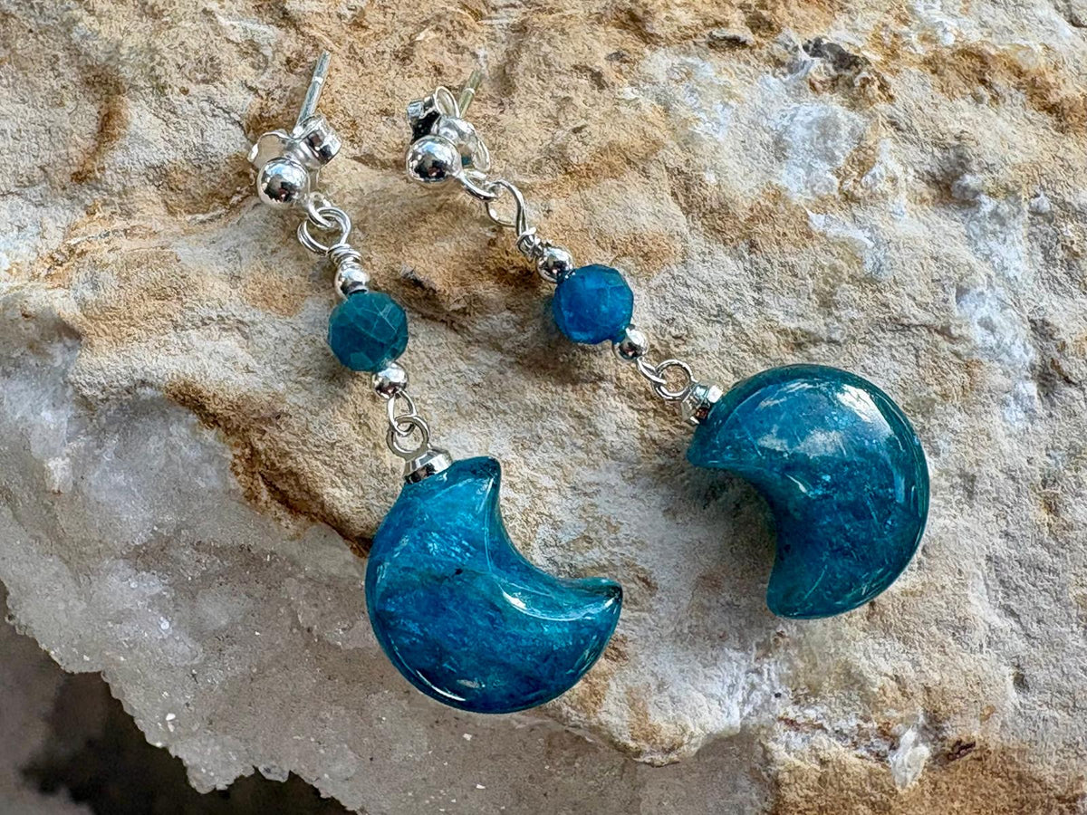 A pair of dangle earrings with polished blue crescent moon charms, blue faceted beads, and silver-toned metal hooks, displayed on a textured beige rock surface.