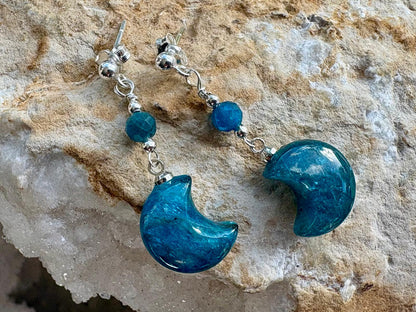 A pair of dangle earrings with polished blue crescent moon charms, blue faceted beads, and silver-toned metal hooks, displayed on a textured beige rock surface.