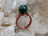 A handmade ring featuring a polished dark green spherical stone set in a copper-colored wire band, displayed on a bed of white crystal formations.