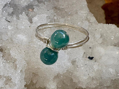 A silver wire-wrapped ring with two smooth, polished teal-green gemstone beads, displayed on a bed of white crystal formations.