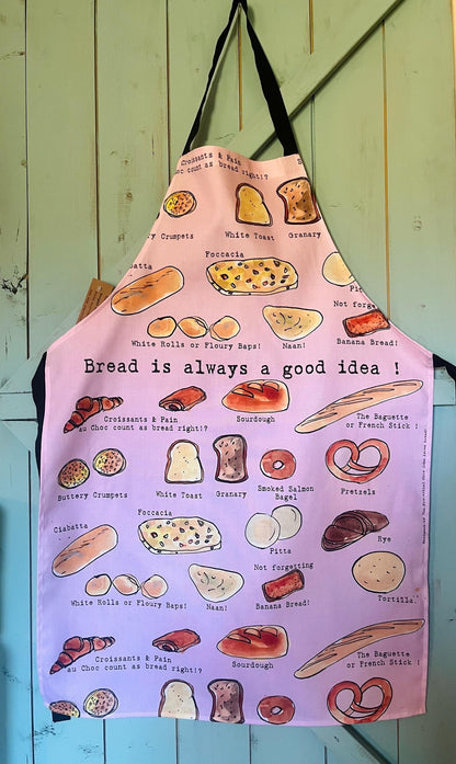A pink kitchen apron with a black neck strap and waist loop, printed with various illustrated breads, croissants, and the text &