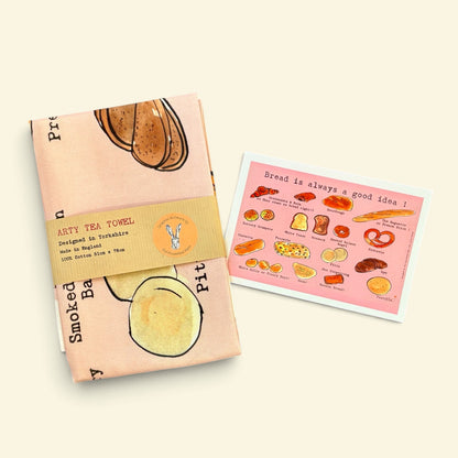 A folded tea towel with a bread-themed print and a tan paper band label, placed next to a matching printed card on a warm cream background.