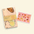 A folded tea towel with a bread-themed print and a tan paper band label, placed next to a matching printed card on a warm cream background.