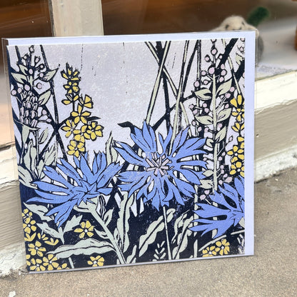A square art print or card featuring a botanical illustration of blue cornflowers and yellow flowers with black linework on a light gray background, photographed leaning against a doorway or shelf.