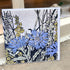 A square art print or card featuring a botanical illustration of blue cornflowers and yellow flowers with black linework on a light gray background, photographed leaning against a doorway or shelf.