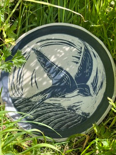 A round ceramic plate with a navy blue and white linocut-style print of a flying bird, photographed outdoors on green grass.