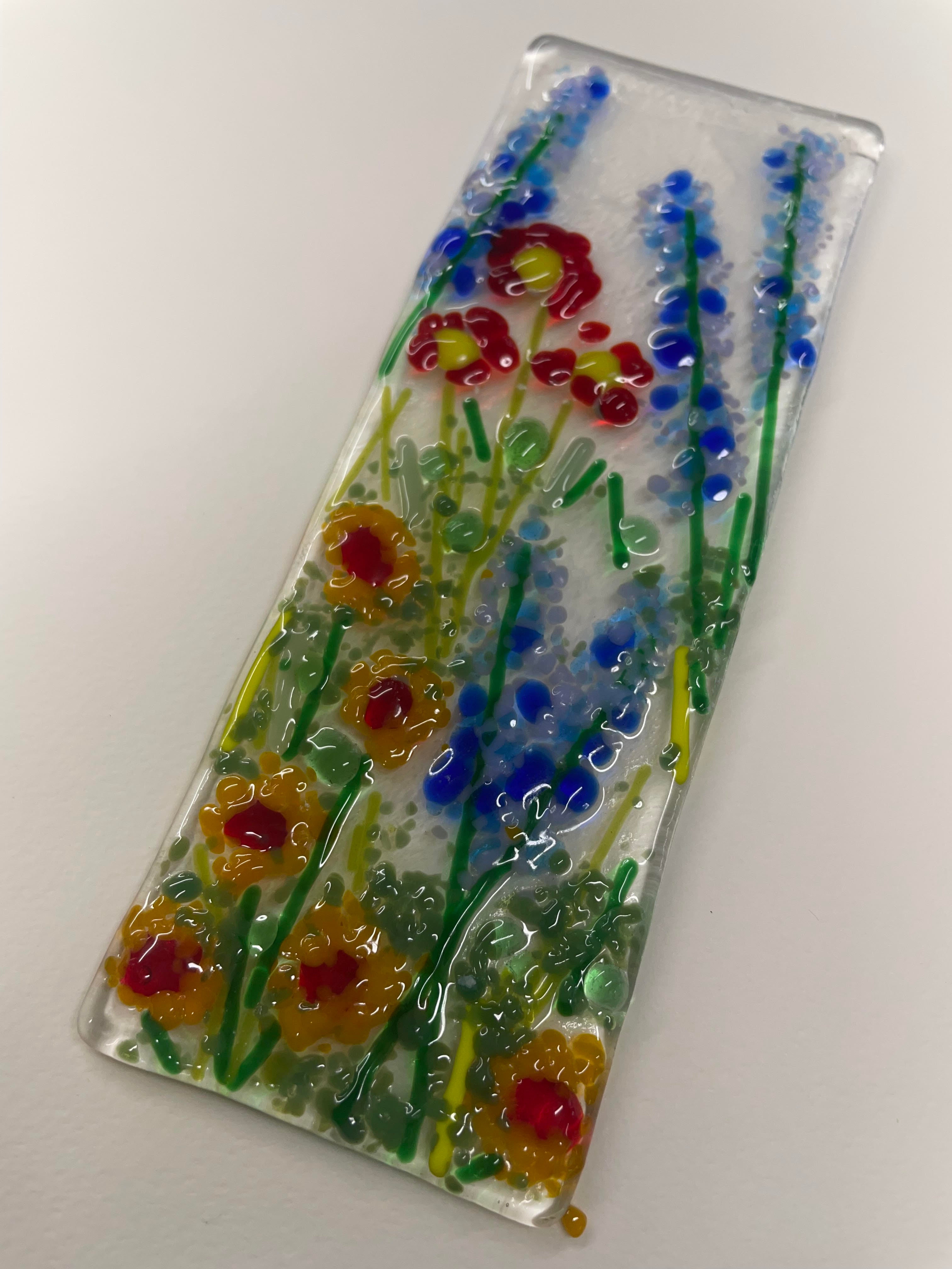 A rectangular clear glass plaque featuring hand-blown floral designs in red, yellow, and blue with green stems, displayed diagonally on a plain white background.
