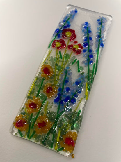 A rectangular clear glass plaque featuring hand-blown floral designs in red, yellow, and blue with green stems, displayed diagonally on a plain white background.
