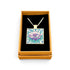 A square pendant necklace displayed inside an open tan gift box with a black velvet insert. The pendant features a floral design with purple flowers, pink blossoms, and green leaves on a light blue background, suspended from a silver-toned chain.