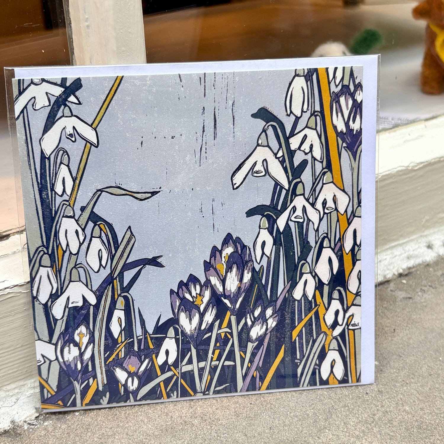 A square art card featuring a stylized floral illustration of snowdrops and crocuses in white, purple, navy, gray, and mustard yellow, leaning against a window ledge.