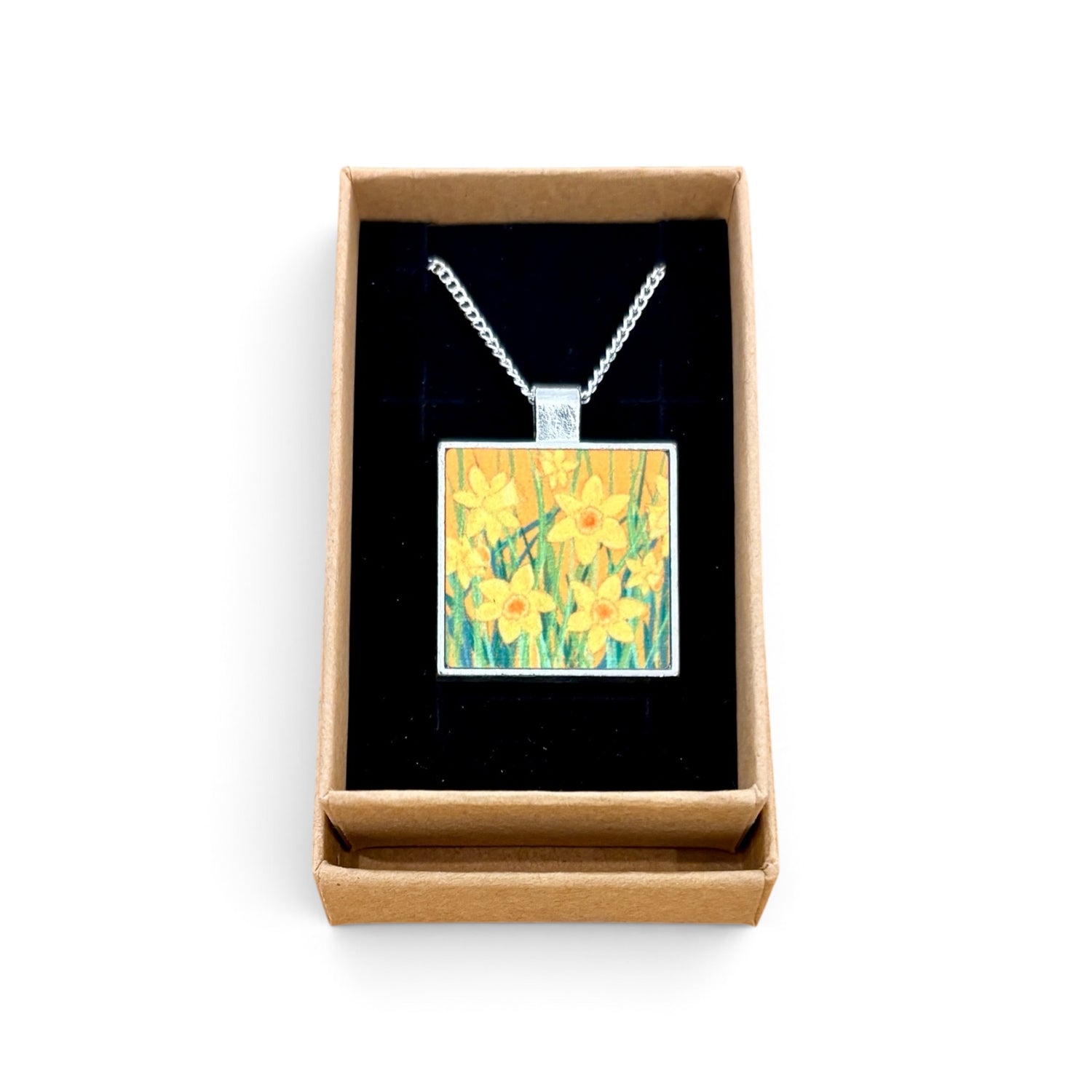 A square pendant necklace with a daffodil painting design, displayed inside an open brown gift box with a black velvet lining on a white background.