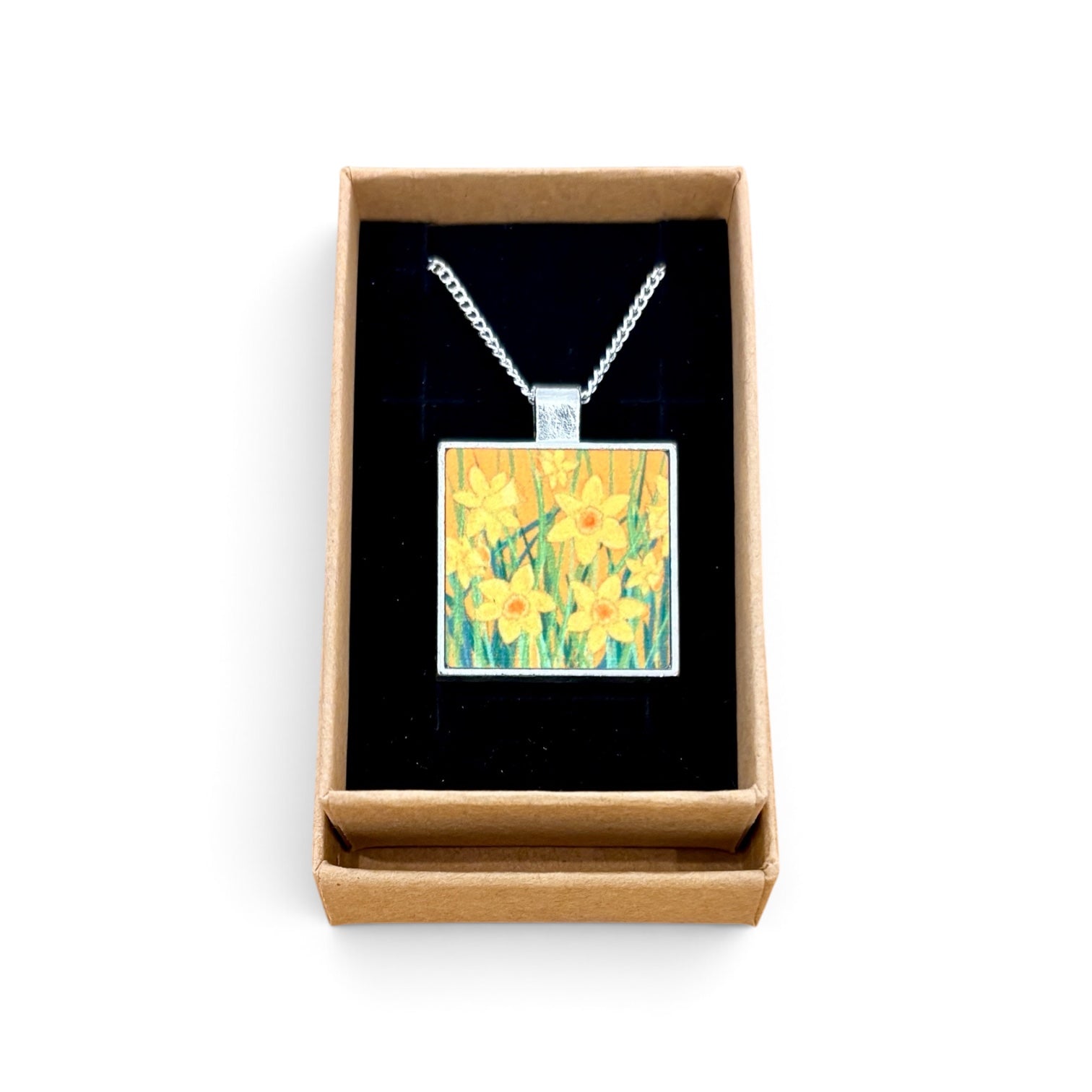 A square pendant necklace with a daffodil painting design, displayed inside an open brown gift box with a black velvet lining on a white background.