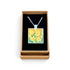 A square pendant necklace with a daffodil painting design, displayed inside an open brown gift box with a black velvet lining on a white background.