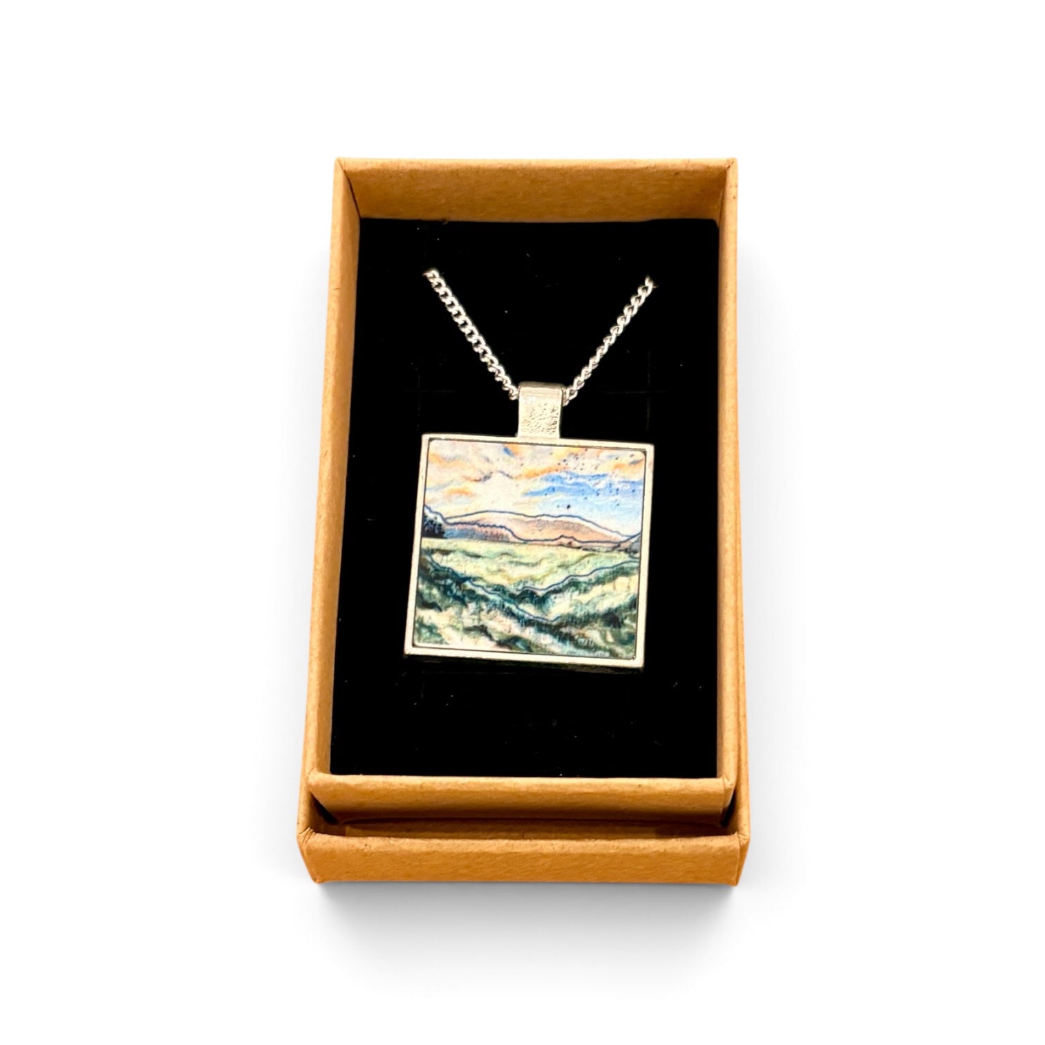A square pendant necklace displayed in an open tan gift box with a black interior. The pendant features a colorful landscape painting with blue, green, yellow, and brown tones, suspended from a silver-toned chain.