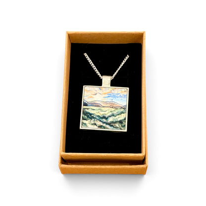A square pendant necklace displayed in an open tan gift box with a black interior. The pendant features a colorful landscape painting with blue, green, yellow, and brown tones, suspended from a silver-toned chain.