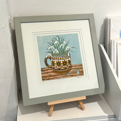A framed art print featuring a colorful illustration of white calla lilies growing out of a patterned ceramic mug, displayed on a small wooden easel in a gallery setting.