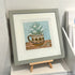 A framed art print featuring a colorful illustration of white calla lilies growing out of a patterned ceramic mug, displayed on a small wooden easel in a gallery setting.