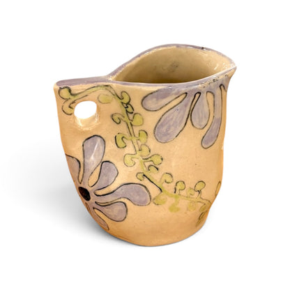 A small handmade ceramic pitcher with a warm beige body, decorated with hand-painted lavender-purple floral motifs and olive-green vines, set against a plain white background.