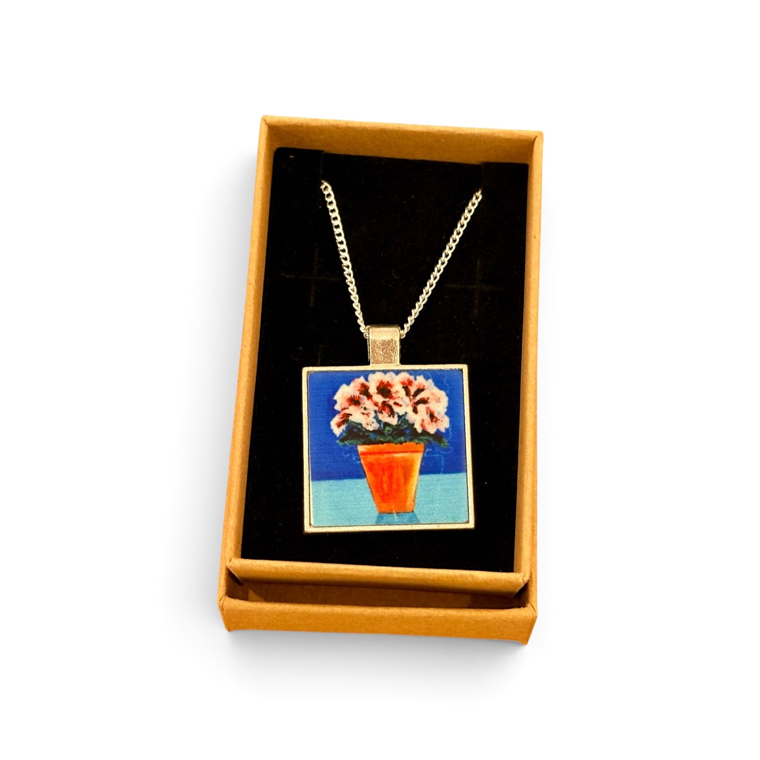 A gold-tone chain necklace with a square pendant featuring a colorful painting of pink flowers in an orange pot, displayed inside an open tan gift box with a black velvet insert on a white background.