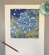 A square art print featuring blue flowers and green leaves on a dark navy background, displayed on a white sheet of paper with a pencil and a glass dish nearby.