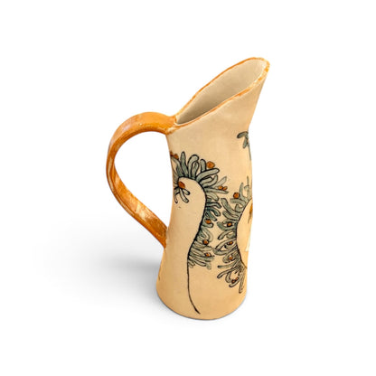 A handmade ceramic pitcher with a warm beige body, a glossy orange-brown handle and rim, and a hand-painted green floral pattern on the side, set against a plain white background.
