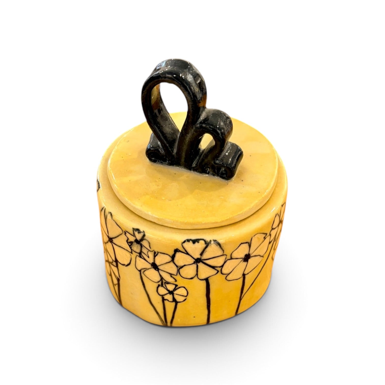 A small round yellow ceramic lidded container with black floral line drawings on the side and a glossy black loop handle on top, shown on a white background.