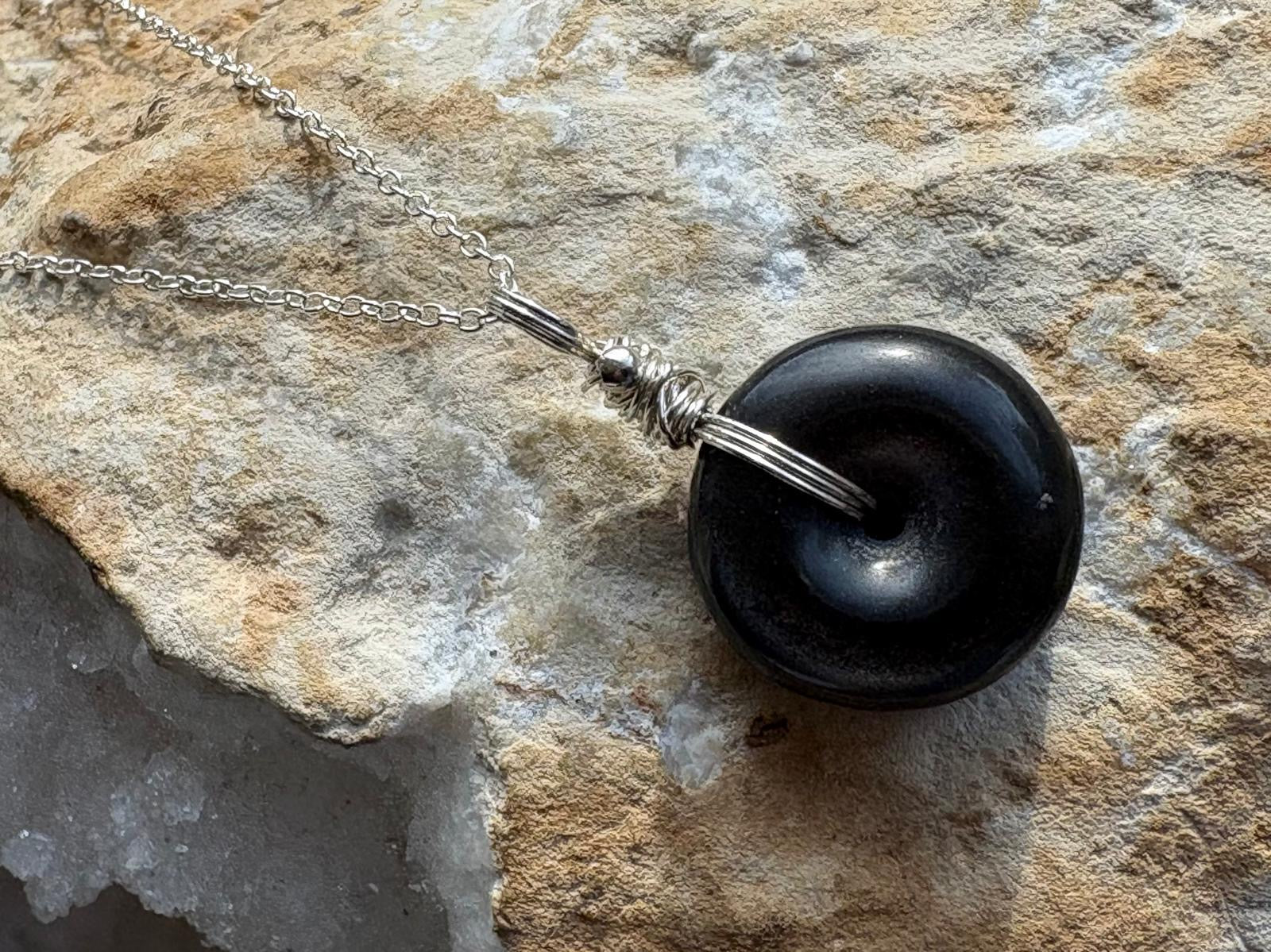 A black circular pendant necklace with a silver chain and wrapped bail, displayed on a textured beige rock surface.