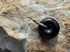 A black circular pendant necklace with a silver chain and wrapped bail, displayed on a textured beige rock surface.