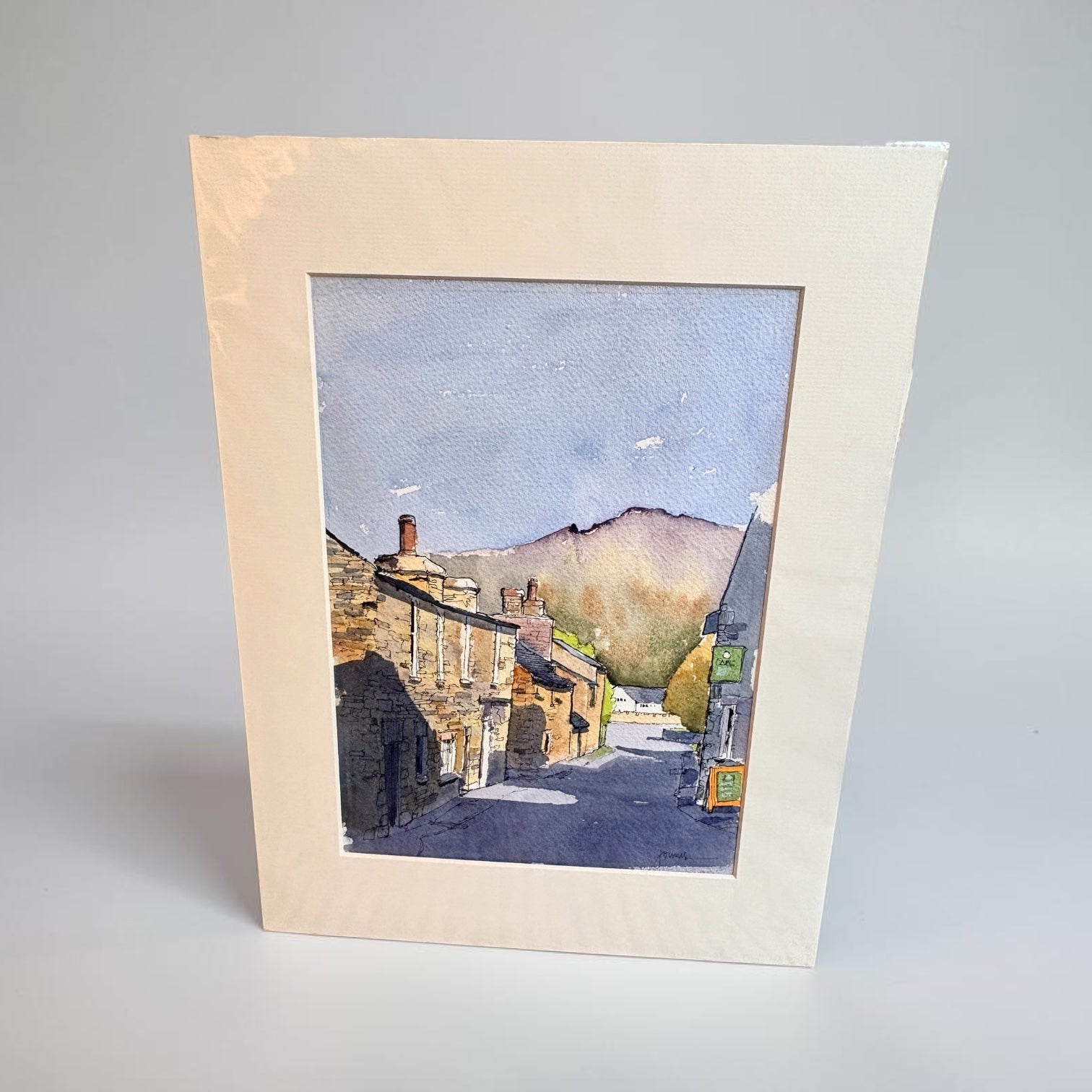 A square watercolor art print depicting a quaint village street with stone buildings, chimneys, and distant hills under a blue sky, mounted with a wide cream-colored mount on a neutral light gray background.