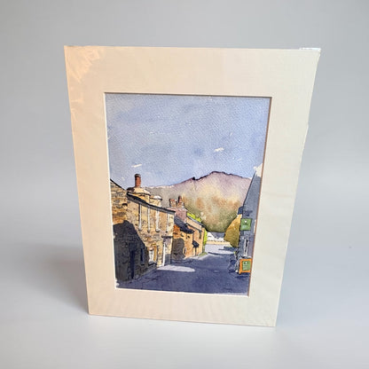 A square watercolor art print depicting a quaint village street with stone buildings, chimneys, and distant hills under a blue sky, mounted with a wide cream-colored mount on a neutral light gray background.