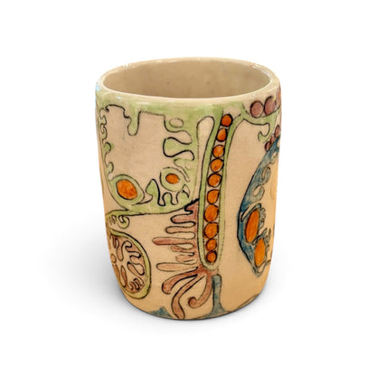 A handmade ceramic cup with a beige base, decorated with organic, abstract patterns in light green, orange, and blue glazes, shown on a plain white background.