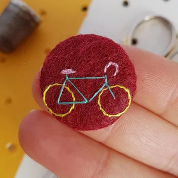 A close-up of a hand holding a small circular felt patch featuring a stylized bicycle embroidered in light blue, yellow, and pink on a dark red background.