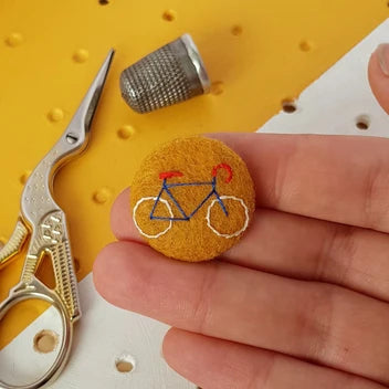 A hand holds a small round felt pin featuring an embroidered bicycle design in blue, red, and white. In the background, gold scissors and a metal thimble rest on a yellow and white pegboard surface.