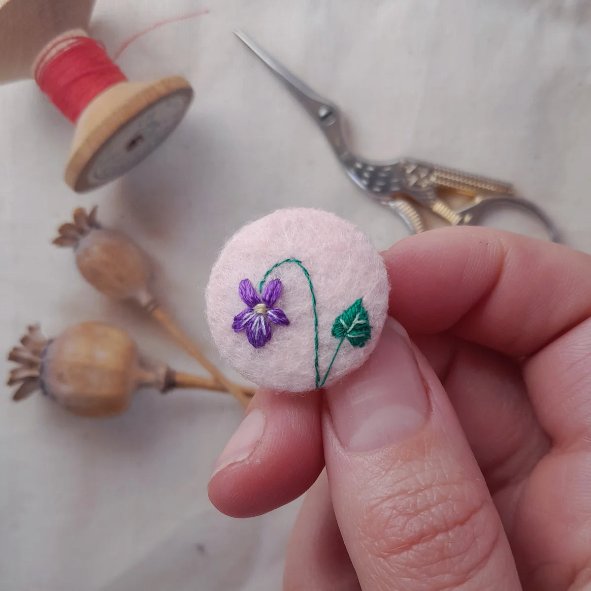 A hand holds a small round felt pin featuring an embroidered purple flower with a green stem and leaf. The background is softly blurred with sewing tools, including a spool of red thread, scissors, and wooden poppy pods.