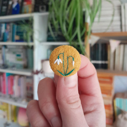 A hand holding a small circular felt pin featuring embroidered white snowdrops and green stems, with a blurred background of bookshelves and a plant.