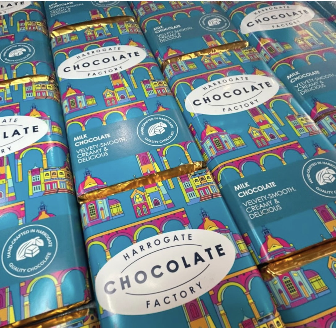 Multiple bars of Harrogate Chocolate Factory chocolate arranged in a grid, wrapped in bright turquoise paper with a colorful architectural illustration and gold foil edges.