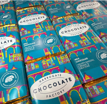 Multiple bars of Harrogate Chocolate Factory chocolate arranged in a grid, wrapped in bright turquoise paper with a colorful architectural illustration and gold foil edges.