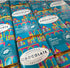 Multiple bars of Harrogate Chocolate Factory chocolate arranged in a grid, wrapped in bright turquoise paper with a colorful architectural illustration and gold foil edges.
