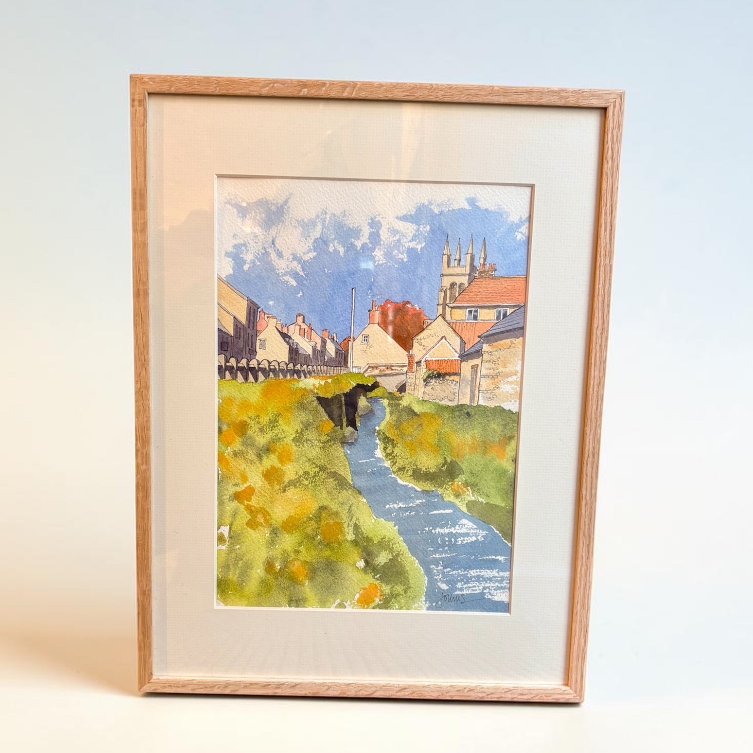 A framed watercolor painting of a village scene with a blue stream, green grassy banks, and buildings under a cloudy sky, set in a light wood frame with a cream mount.